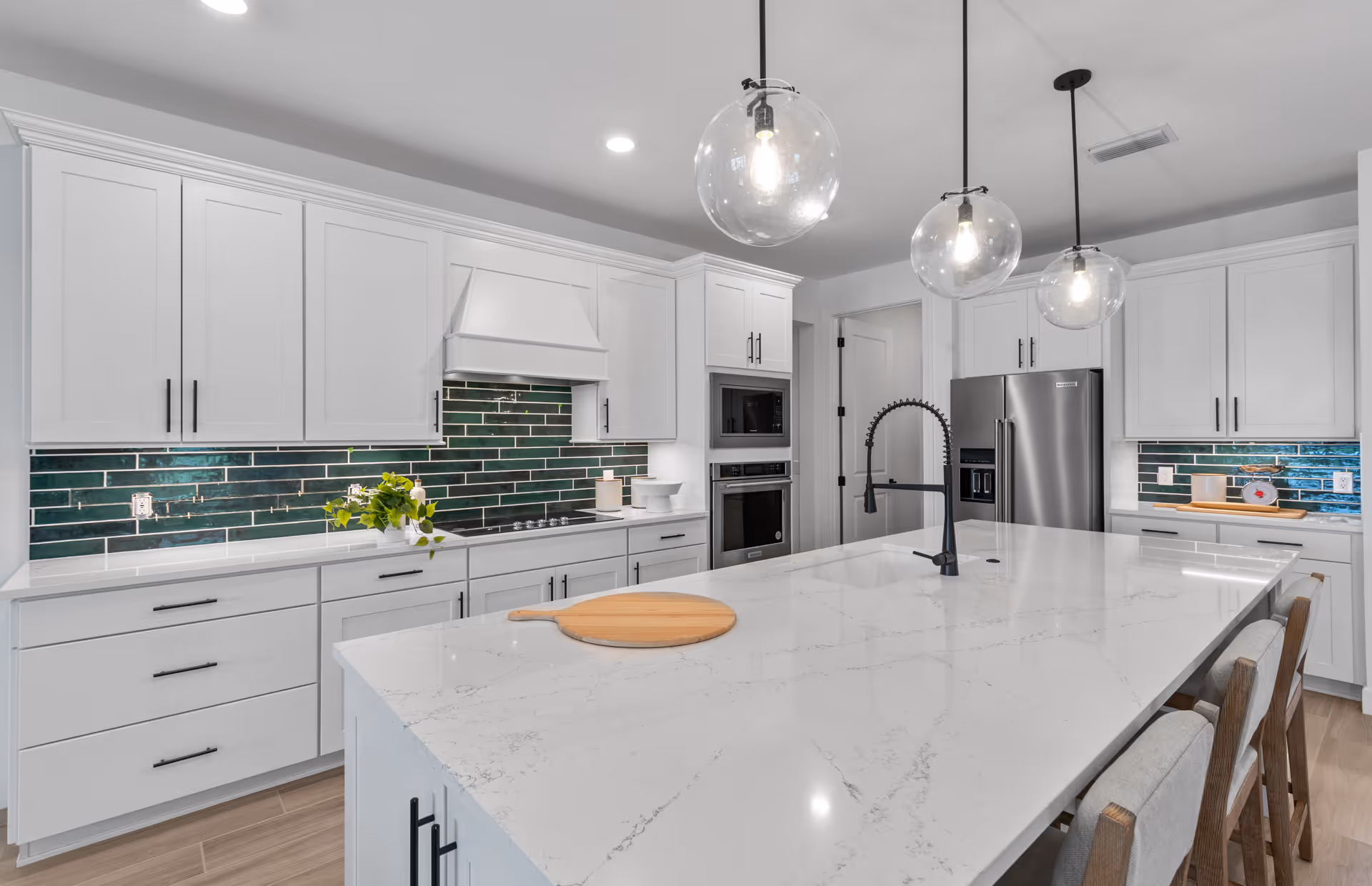 Modern kitchen with white cabinetry, a large marble island countertop with a built-in sink and black faucet, three pendant lights with clear glass globes hanging above the island, stainless steel refrigerator, built-in microwave and oven, and a green tile backsplash.