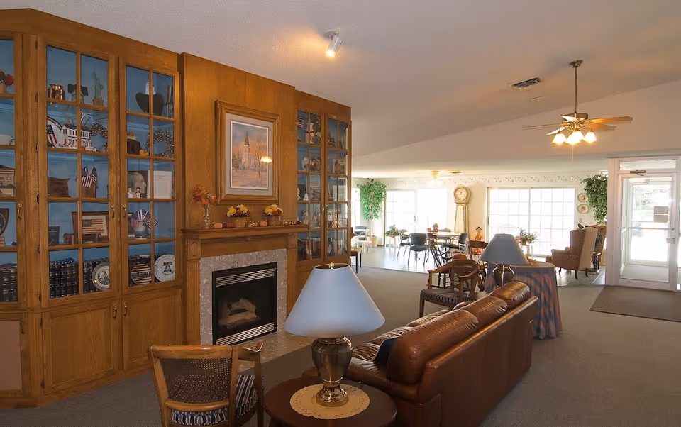 A cozy senior living facility common area featuring a brown leather sofa, wooden chairs, a fireplace with a wooden mantel, and built-in wooden shelves with decorative items. In the background, there is a dining area with tables and chairs, large windows letting in natural light, a grandfather clock, and plants near the entrance door.