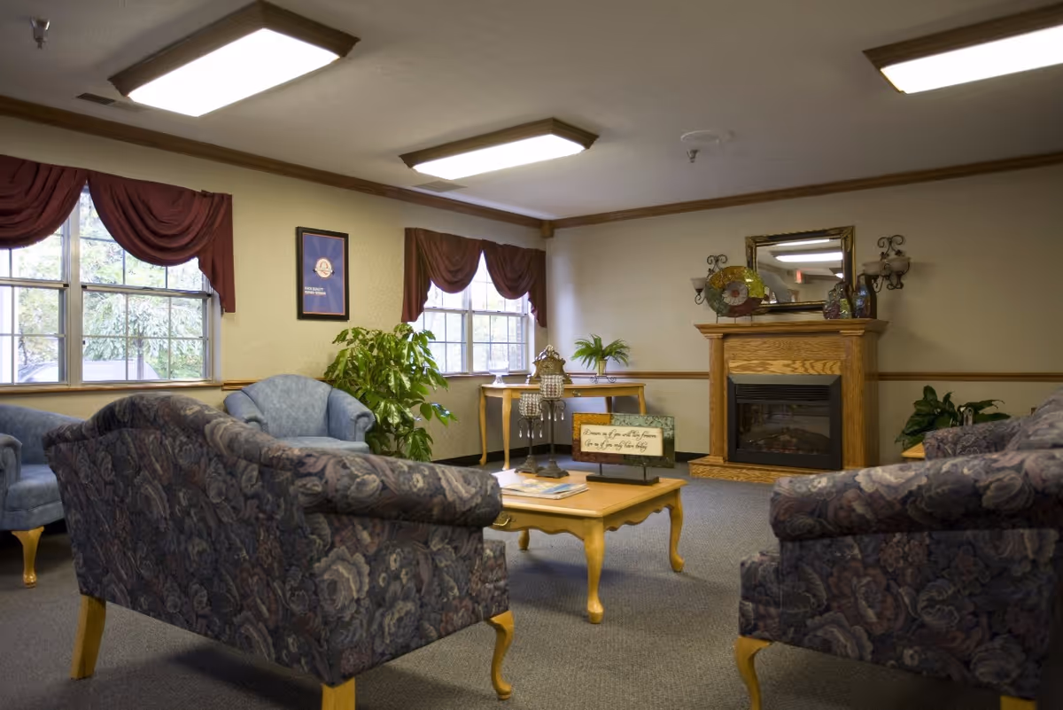 A cozy living room area with floral patterned sofas and blue armchairs arranged around a wooden coffee table. The room features large windows with burgundy curtains, a wooden fireplace with decorative items on the mantel, a mirror above the fireplace, and indoor plants near the windows and fireplace.
