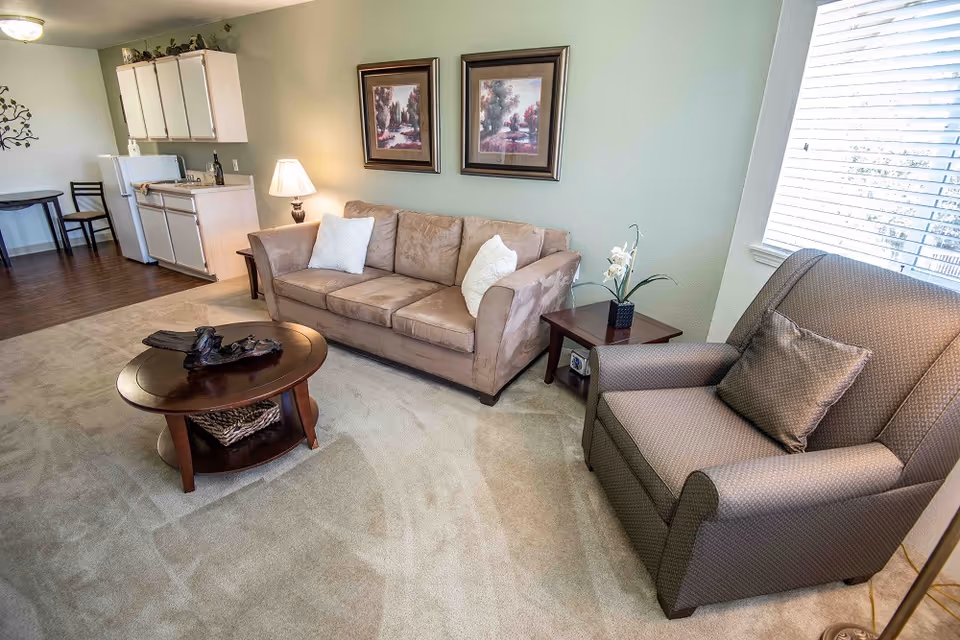 A cozy living room area in a senior living facility featuring a beige sofa with two white pillows, a patterned armchair with a matching pillow, a wooden coffee table with decorative items, and a side table with a lamp and a small plant. The room has light green walls with two framed landscape paintings and a large window with blinds letting in natural light. In the background, there is a small kitchenette with white cabinets, a refrigerator, a sink, and a small dining table with two chairs.