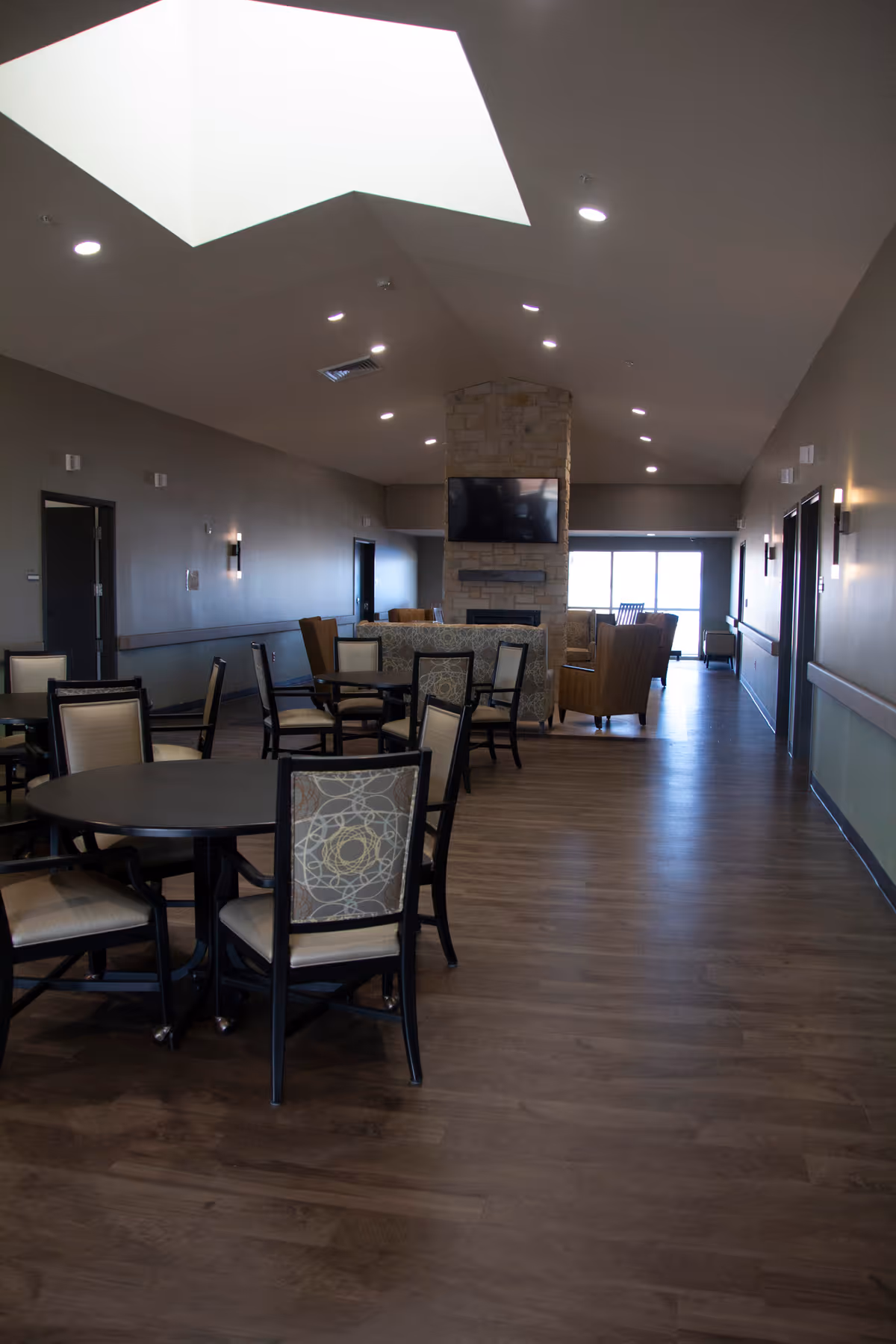 Spacious common room with round dining tables and chairs leading to a seating area with a stone fireplace and wall-mounted TV.