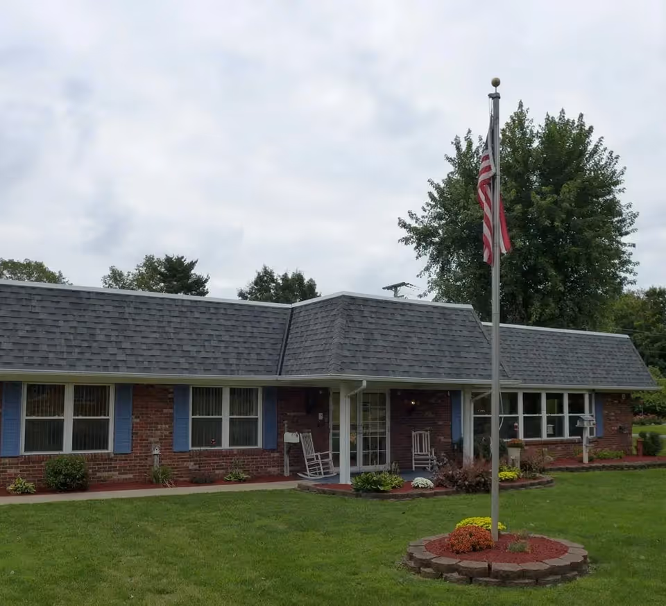 Single-story brick building with a gray shingled roof, blue window shutters, and a covered entrance with two white rocking chairs. In front of the building is a flagpole with an American flag and a circular flower bed at its base. The lawn is green and well-maintained, with trees in the background under a cloudy sky.