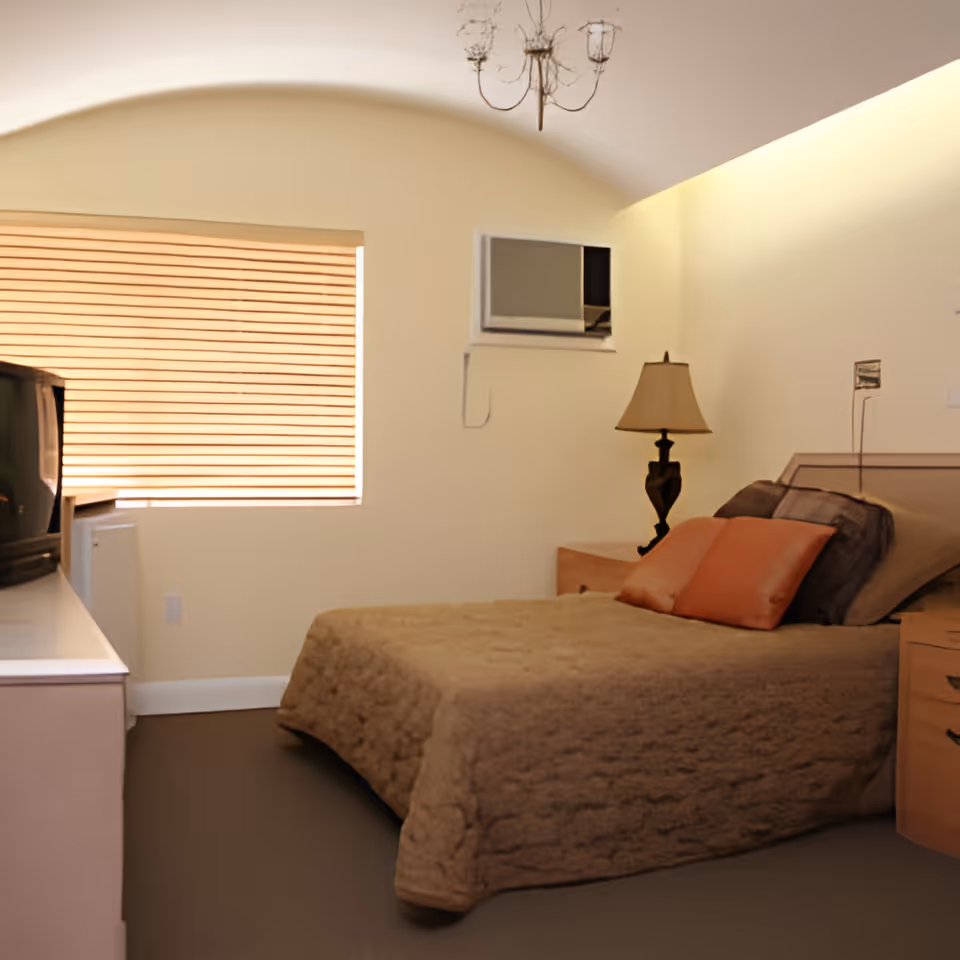 A cozy bedroom with a bed covered in a beige quilt and several pillows, a wooden nightstand with a lamp, a window with closed blinds, an air conditioning unit mounted on the wall, and a TV on a white dresser.