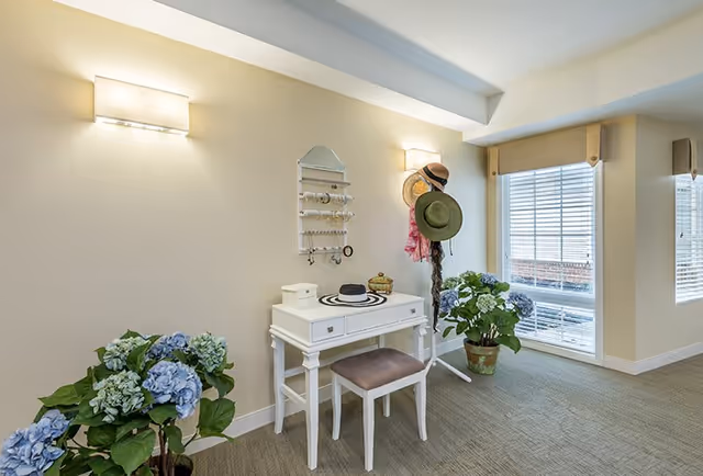 A bright interior corner featuring a white vanity table with a cushioned stool, a hat and jewelry organizer mounted on the wall, and a coat rack holding hats and scarves. Two potted plants with blue flowers are placed on either side of the vanity. Large windows with blinds allow natural light to fill the space.