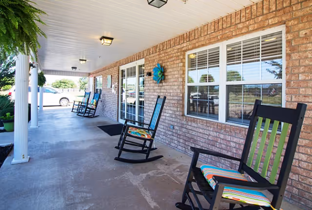 Covered porch area with brick walls and several black rocking chairs with colorful cushions lined up along the wall. White columns support the roof, and there are windows and a glass door along the brick wall. The porch overlooks a parking area and some greenery.