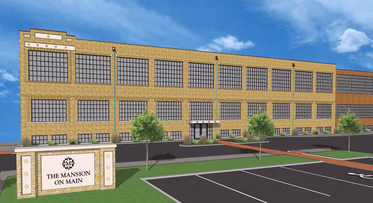 Exterior view of a large, multi-story brick building with many windows under a blue sky. In front of the building is a parking lot and a sign that reads 'The Mansion on Main' with a decorative emblem above the text. There are a few small trees and landscaped areas near the building entrance.