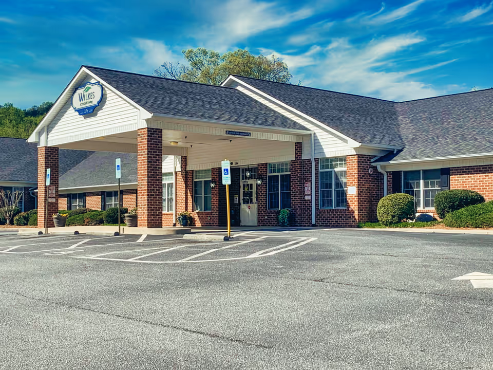 Exterior front view of Wilkes Assisted Living facility showing a brick building with a covered entrance, handicap parking spaces, and a clear blue sky.