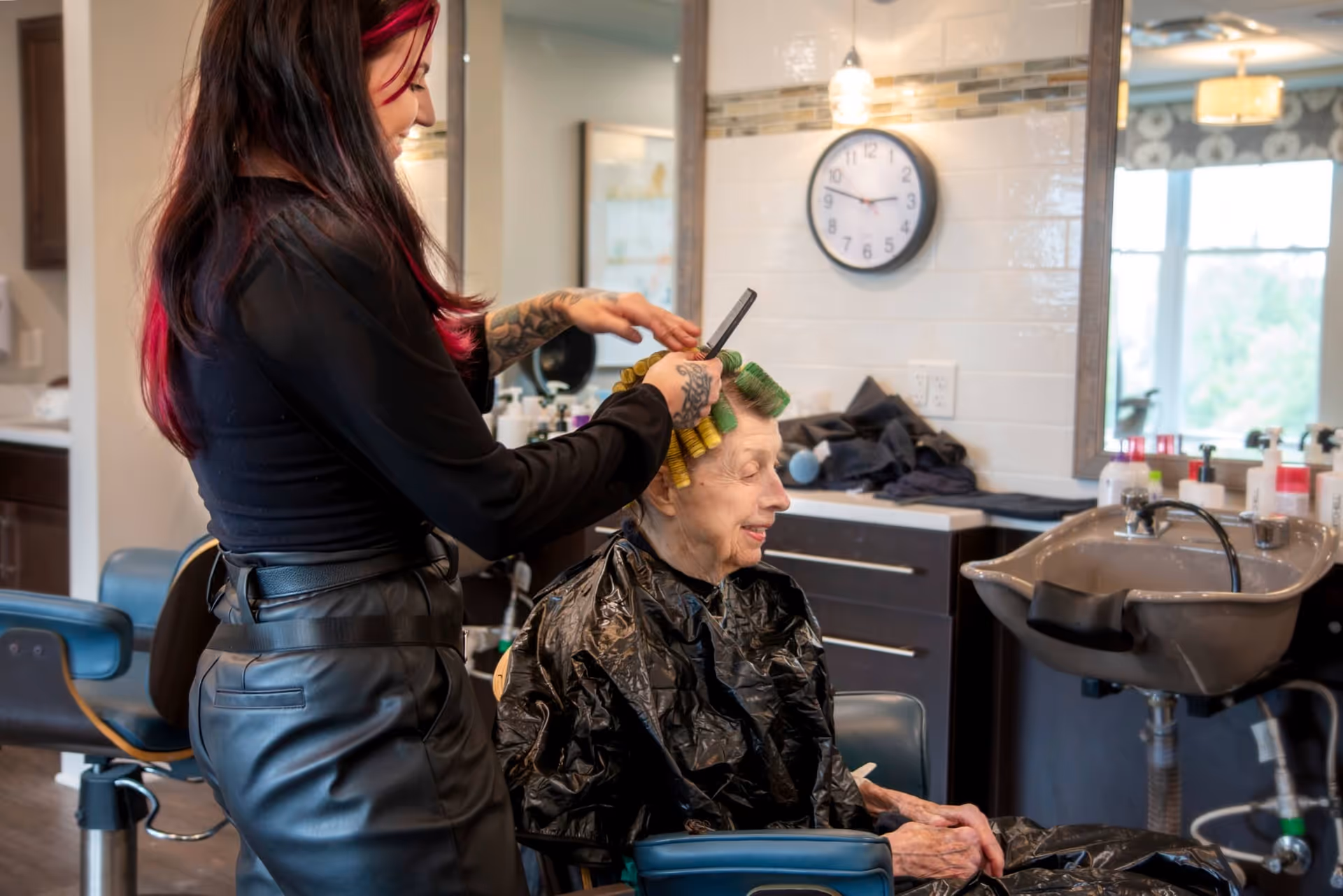 A hairstylist with long dark hair and red highlights is styling an elderly woman's hair with curlers in a salon setting. The elderly woman is seated in a blue salon chair wearing a black cape. The background shows a sink, countertop with hair products, a wall clock, and a large mirror reflecting the room.