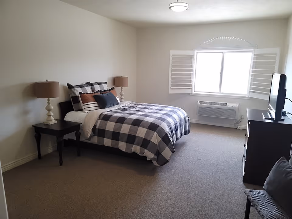 A bright, simply furnished bedroom with a plaid-covered bed, two bedside lamps, a dresser with a TV, and a window with shutters.