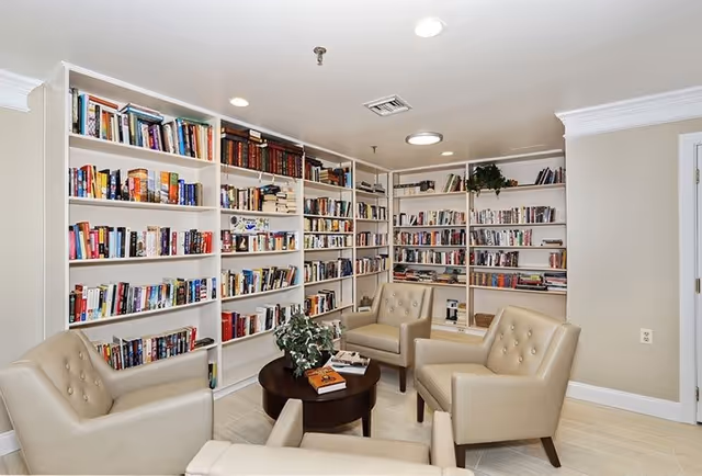 A cozy reading room with beige walls and ceiling lights, featuring four beige leather armchairs arranged around a small round wooden table with books and a plant on it. The walls are lined with white built-in bookshelves filled with a variety of books.