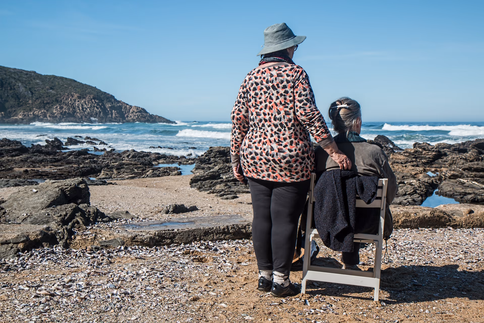 Two people on a rocky beach, one standing with a hand on the other's shoulder while the other sits facing the ocean.