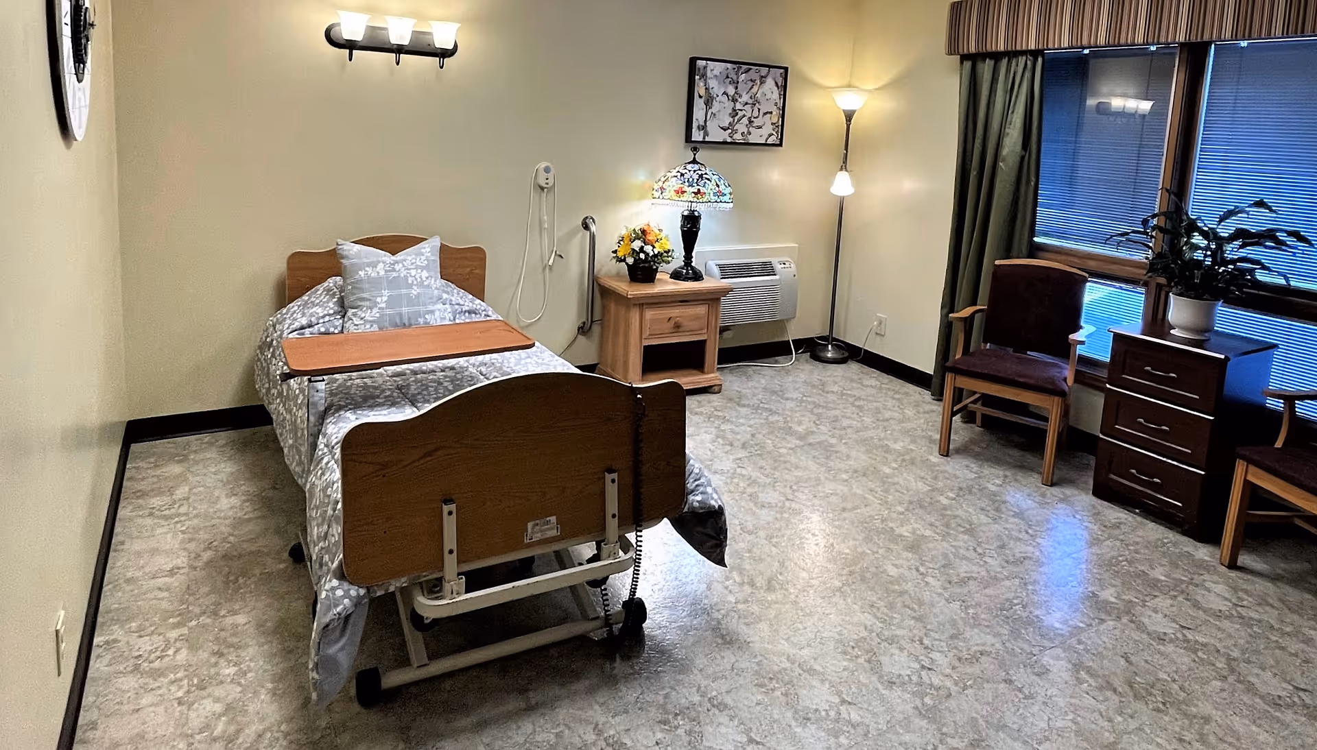 Spacious senior bedroom with a single adjustable hospital-style bed, nightstand, chairs and a window with blinds.