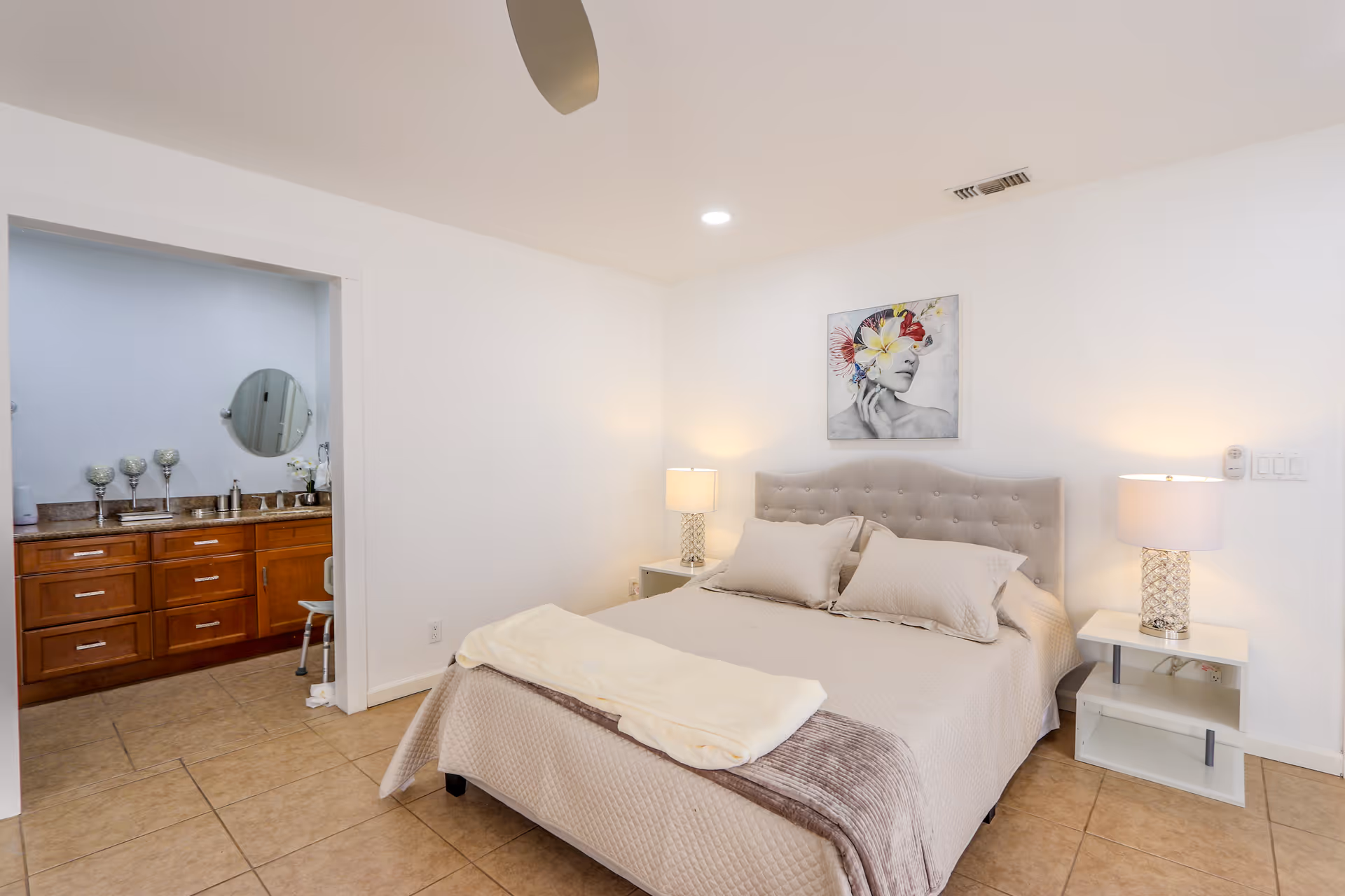 A bedroom with a neatly made bed featuring beige bedding and pillows. There are two bedside tables with lamps on either side of the bed. Above the bed hangs a colorful artwork of a woman's face with flowers and butterflies. To the left, an open doorway leads to a bathroom with a wooden vanity, granite countertop, and a round mirror.