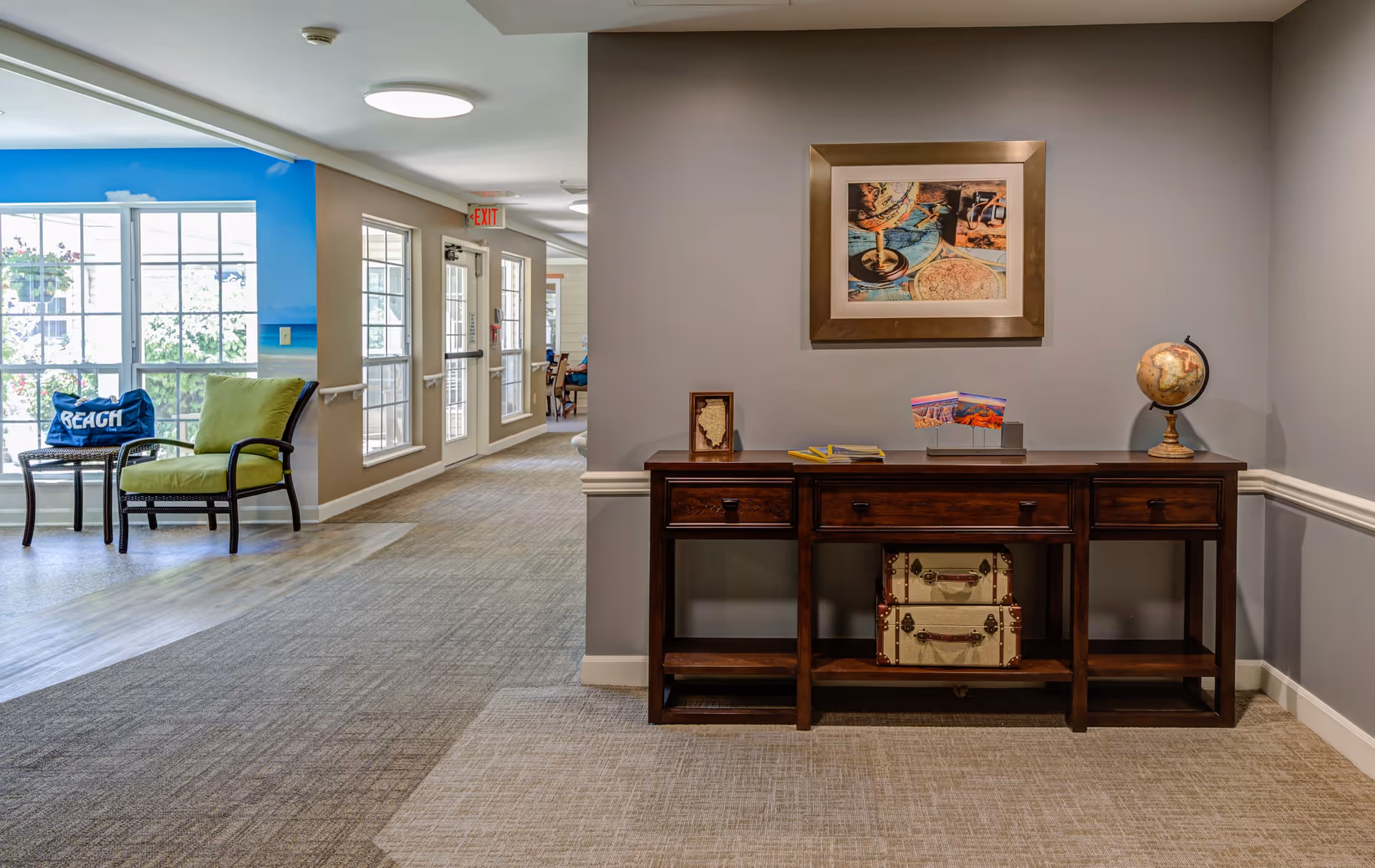 A hallway in a senior living facility with a wooden console table against a gray wall. On the table are decorative items including a globe, framed picture, brochures, and two vintage suitcases on the lower shelf. To the left, there are large windows and a seating area with a green cushioned chair and a small table with a blue pillow that says 'BEACH'. The hallway leads to a dining area visible in the background.
