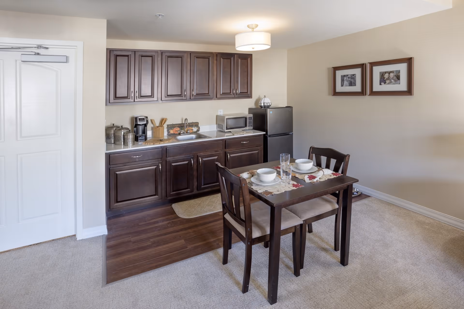 A small kitchen area with dark wood cabinets, a sink, a microwave, a coffee maker, and a mini refrigerator. In front of the kitchen is a dark wood dining table set for two with white bowls, plates, and glasses. The room has beige walls with two framed photos hanging on one wall and a light fixture on the ceiling.