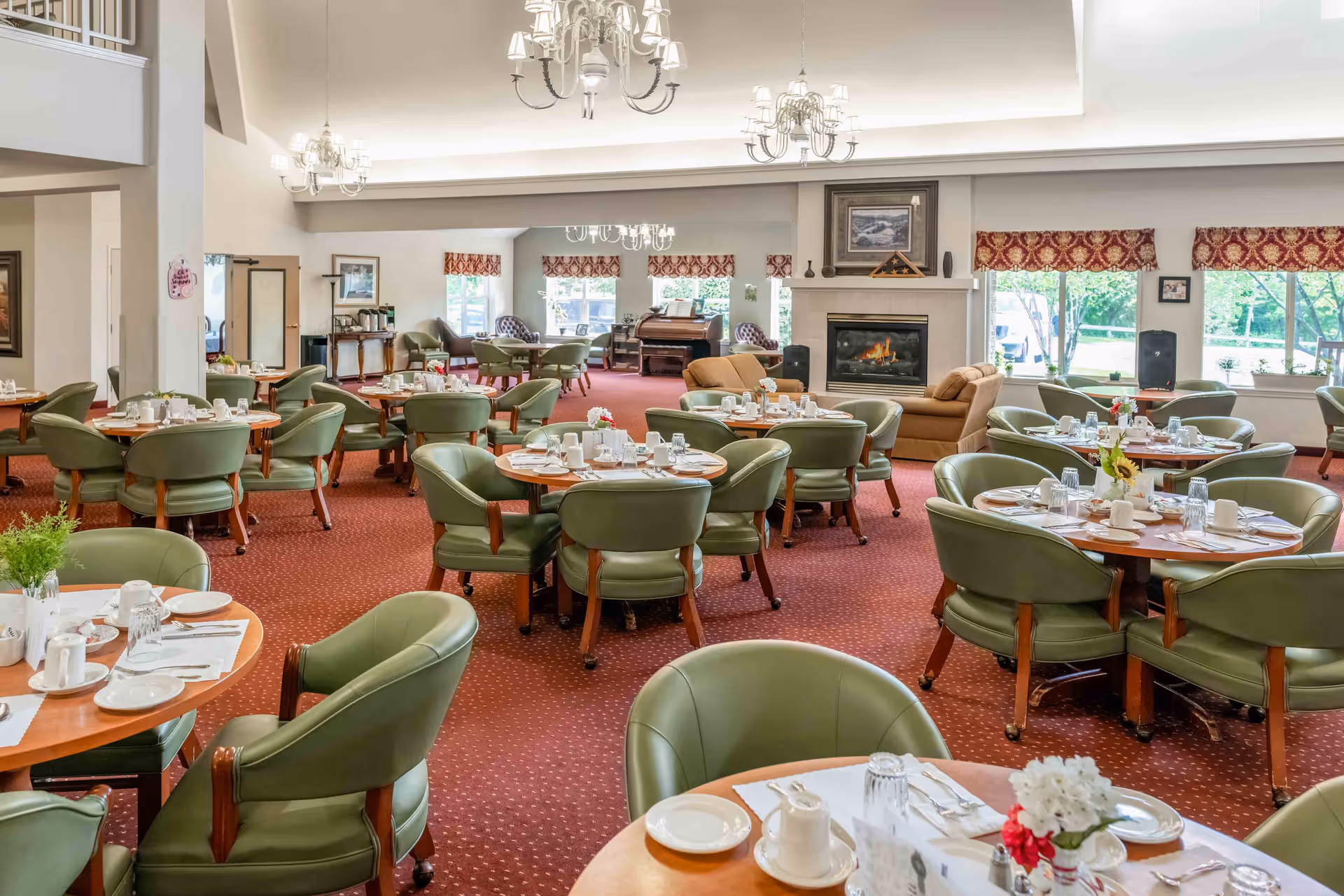 A spacious dining room with round wooden tables set with white plates, cups, and silverware. The room features green cushioned chairs around each table, a red carpeted floor, large windows with red patterned valances, and a fireplace with a lit fire. Chandeliers hang from the ceiling, and there are sofas and additional seating near the fireplace and windows.