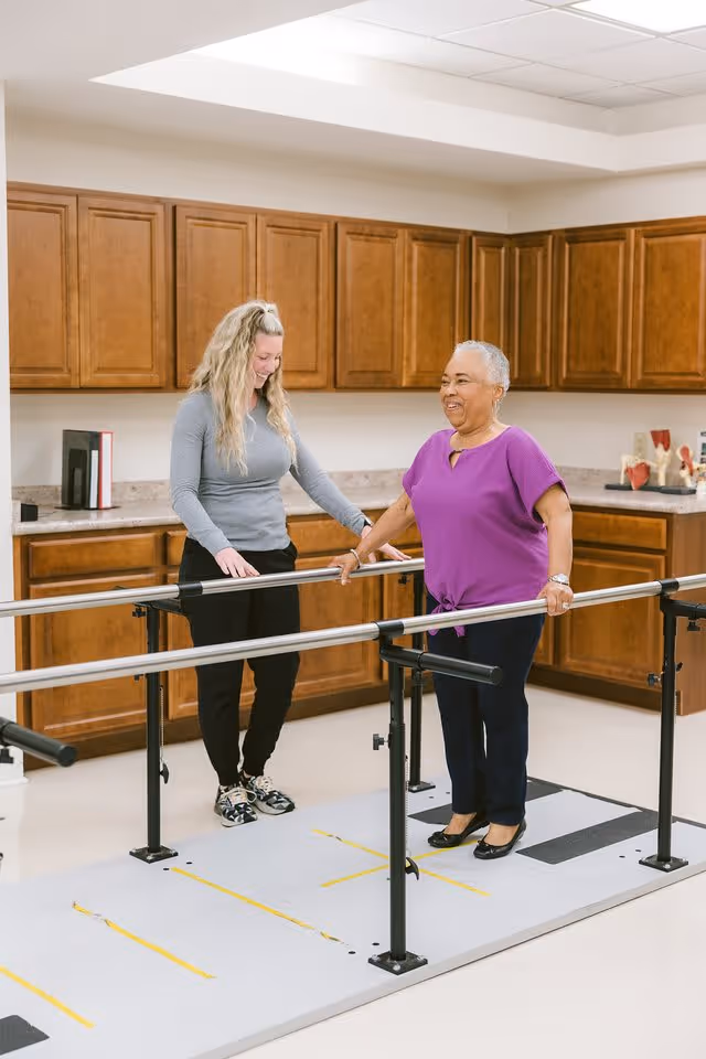 An elderly woman in a purple shirt uses parallel bars for walking exercise in a room with wooden cabinets. A younger woman in a gray long-sleeve shirt and black pants stands beside her, offering support and encouragement.