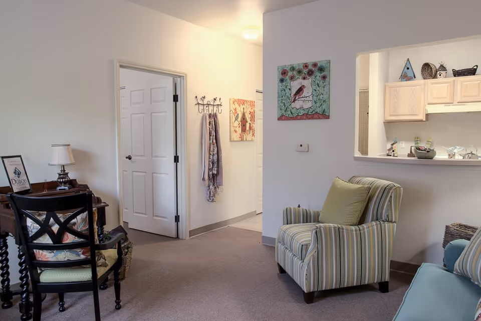 A cozy living area in a senior living facility with a striped armchair and a matching sofa, a small wooden desk with a lamp and framed picture, a wall with colorful bird-themed artwork, and a pass-through window to a kitchen with light wood cabinets.