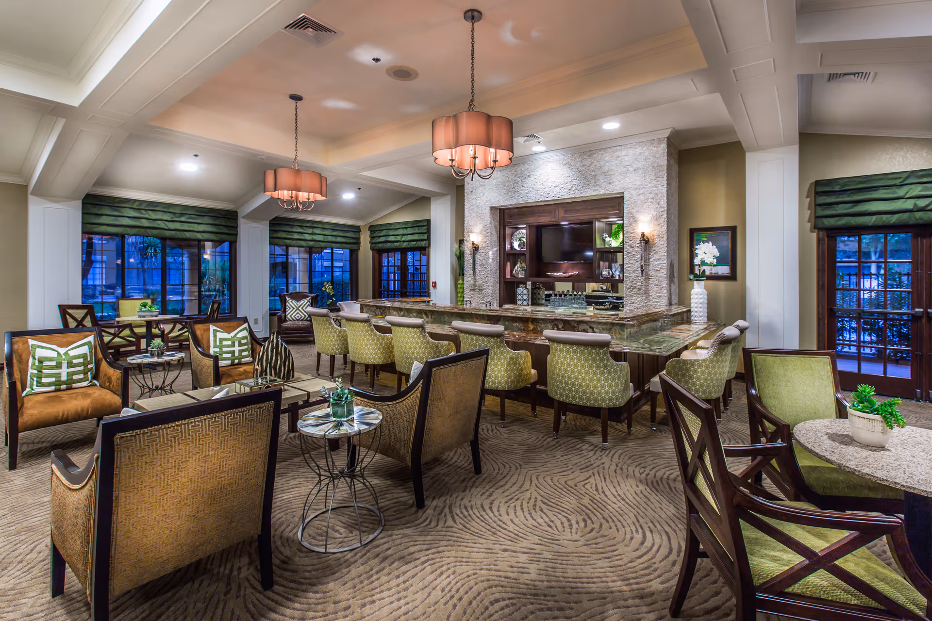 A spacious and elegant common area with a large marble countertop bar surrounded by green patterned chairs. The room features comfortable seating with armchairs and small tables, green window valances, and warm lighting from ceiling fixtures and wall sconces. Large windows and glass doors provide a view of the outside.