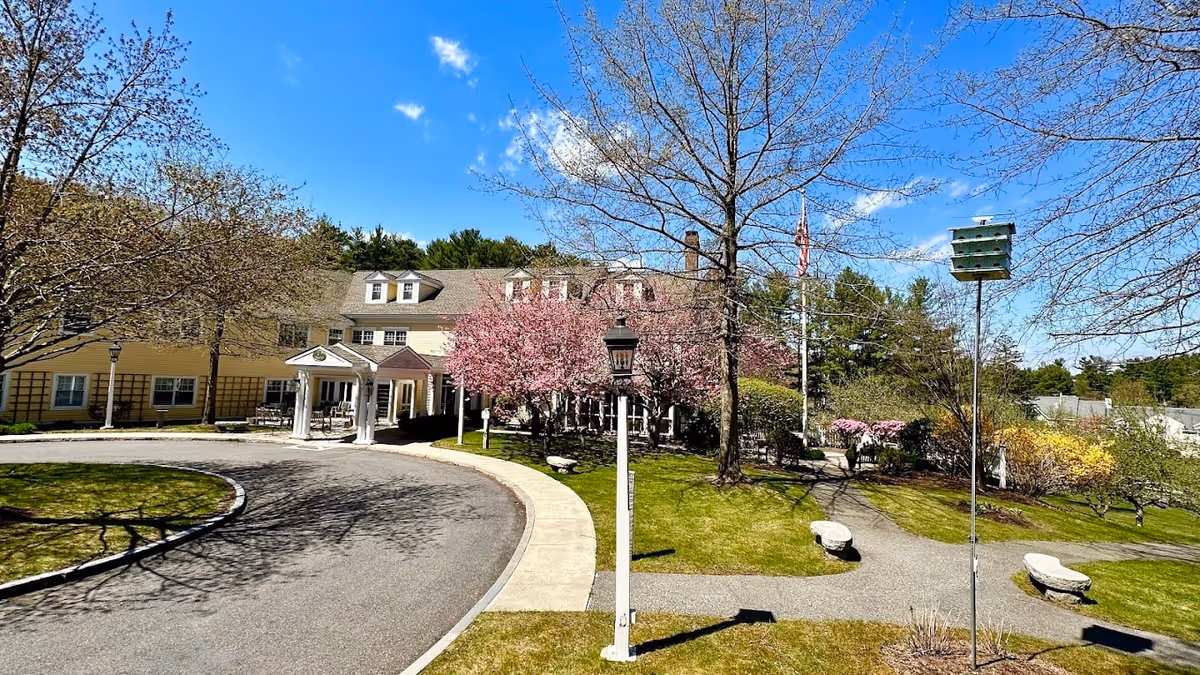 Exterior view of Orchard Hill at Sudbury facility on a clear sunny day with a curved driveway, blooming pink trees, green lawns, and a multi-story building with dormer windows in the background.