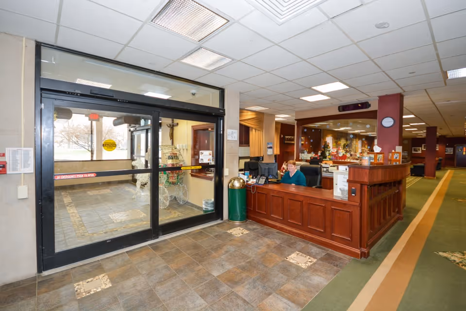 Reception area of Immaculate Mary Center for Rehabilitation & Healthcare with a woman sitting behind a wooden front desk. To the left, there are automatic sliding glass doors leading outside. The floor is tiled near the entrance and carpeted further inside. The ceiling has recessed lighting, and there is a clock on a maroon column behind the desk.