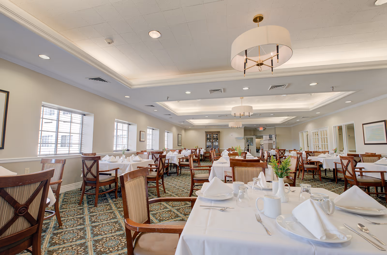 A spacious dining room with multiple tables covered in white tablecloths, each set with white plates, folded napkins, cups, glasses, and silverware. The room has patterned carpet flooring, large windows along one wall, and modern ceiling lights. There are wooden chairs with cushioned seats around the tables, and small flower arrangements on each table.