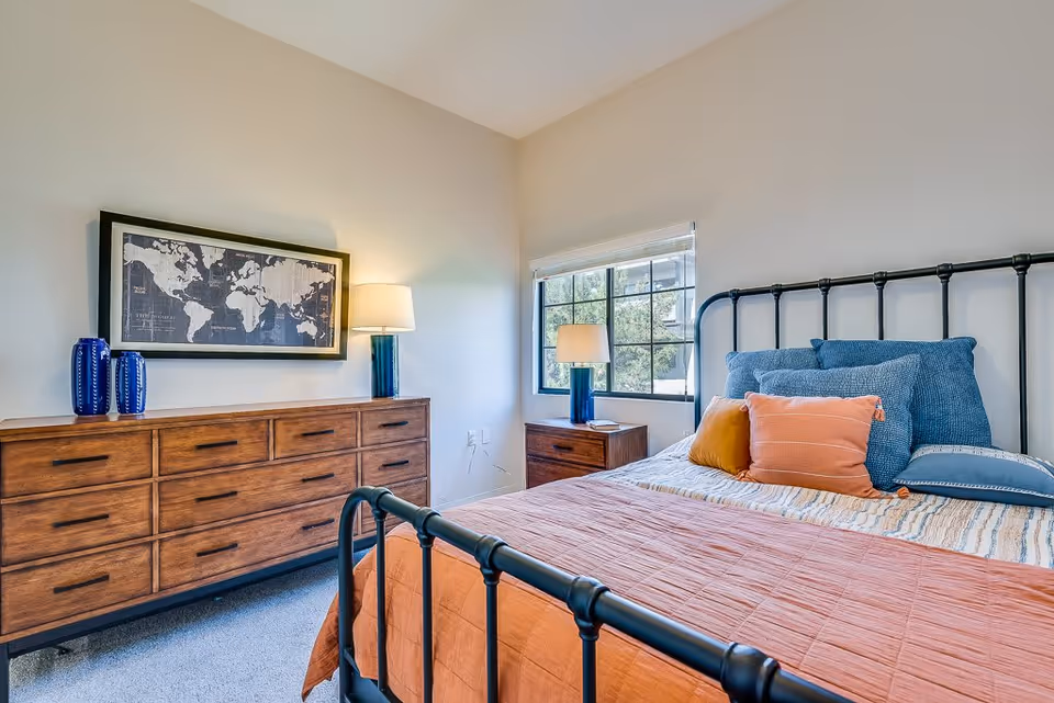 A bright bedroom with a black metal bed frame, a bed made with blue and orange pillows and an orange blanket. There is a wooden dresser with multiple drawers on the left side, decorated with two blue vases and a lamp. Above the dresser hangs a framed world map. A window with white blinds is on the right wall, next to a wooden nightstand with a lamp and a small item on it.