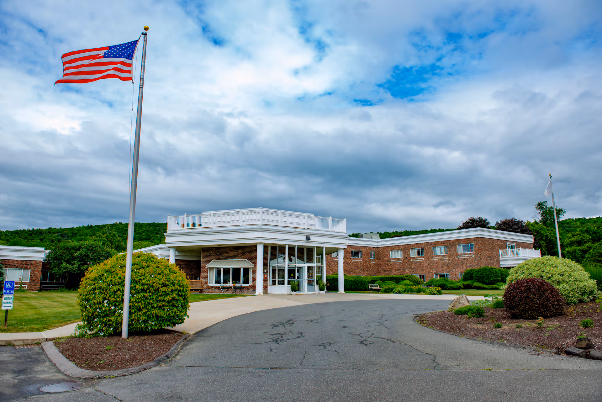 Front exterior of a brick senior living building with an American flag, circular driveway, and landscaped shrubs under a cloudy sky.