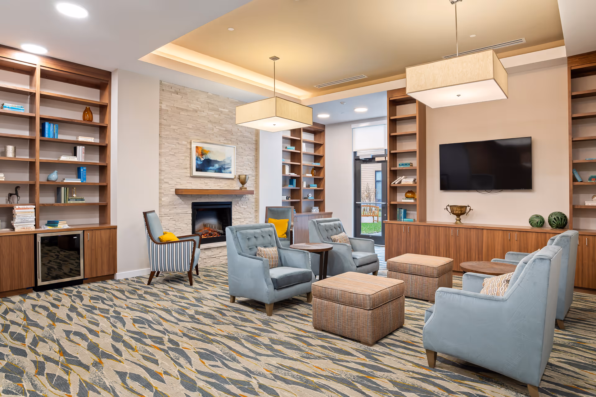 A modern and cozy living room area in a senior living facility featuring light blue upholstered armchairs arranged around two ottomans and small wooden tables. The room has built-in wooden bookshelves with decorative items and books, a wall-mounted flat-screen TV, and a fireplace with a stone surround and a colorful abstract painting above it. The carpet has a patterned design in shades of blue, gray, and beige, and the ceiling has recessed lighting and two large rectangular pendant lights. A glass door leads to an outdoor area with greenery.