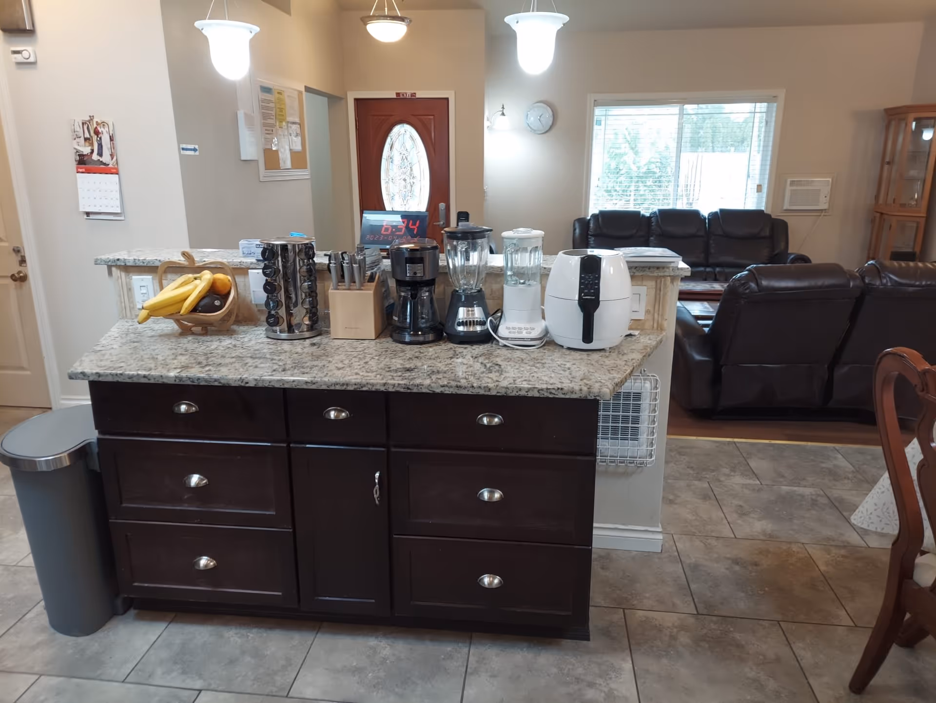 Kitchen island with granite countertop holding appliances (coffee maker, blenders, air fryer) and a fruit bowl, with a living room visible beyond.
