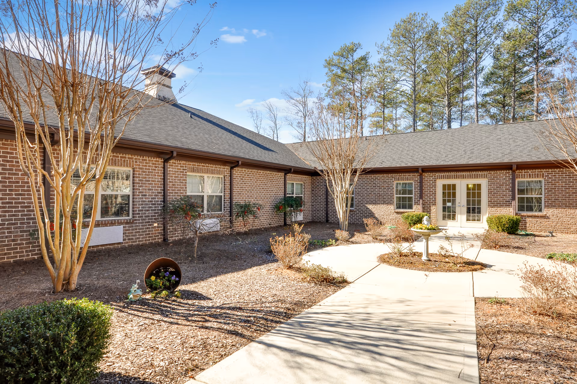 Outdoor courtyard area of a senior living facility with a paved walkway, small garden beds with shrubs and trees, and a brick building with multiple windows and a double door entrance under a clear blue sky.