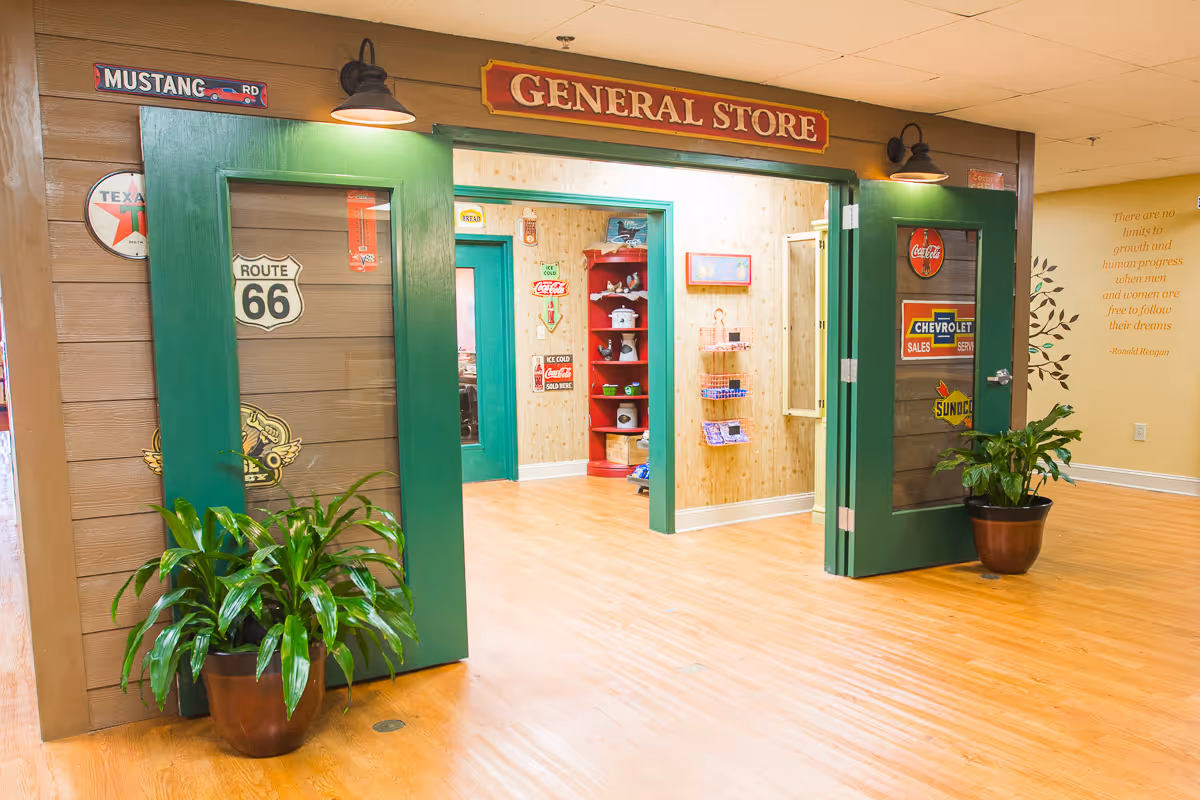 Entrance to a general store inside a facility, featuring green double doors decorated with vintage signs such as Route 66, Coca-Cola, Chevrolet, and Sunoco. There are two potted plants on either side of the entrance and a wooden floor. Inside, shelves with various items are visible, and a wall quote by Ronald Reagan is seen on the right.