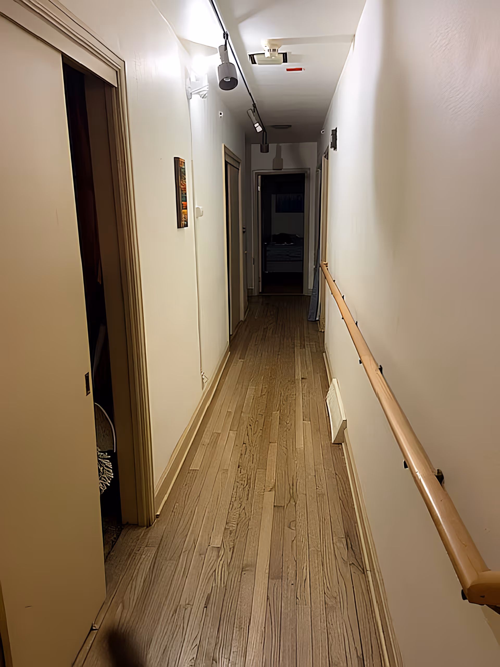 A narrow hallway with wooden flooring and white walls. There are several doorways along the hallway, a wooden handrail on the right wall, and ceiling lights illuminating the space. At the end of the hallway, a room with a bed is partially visible.