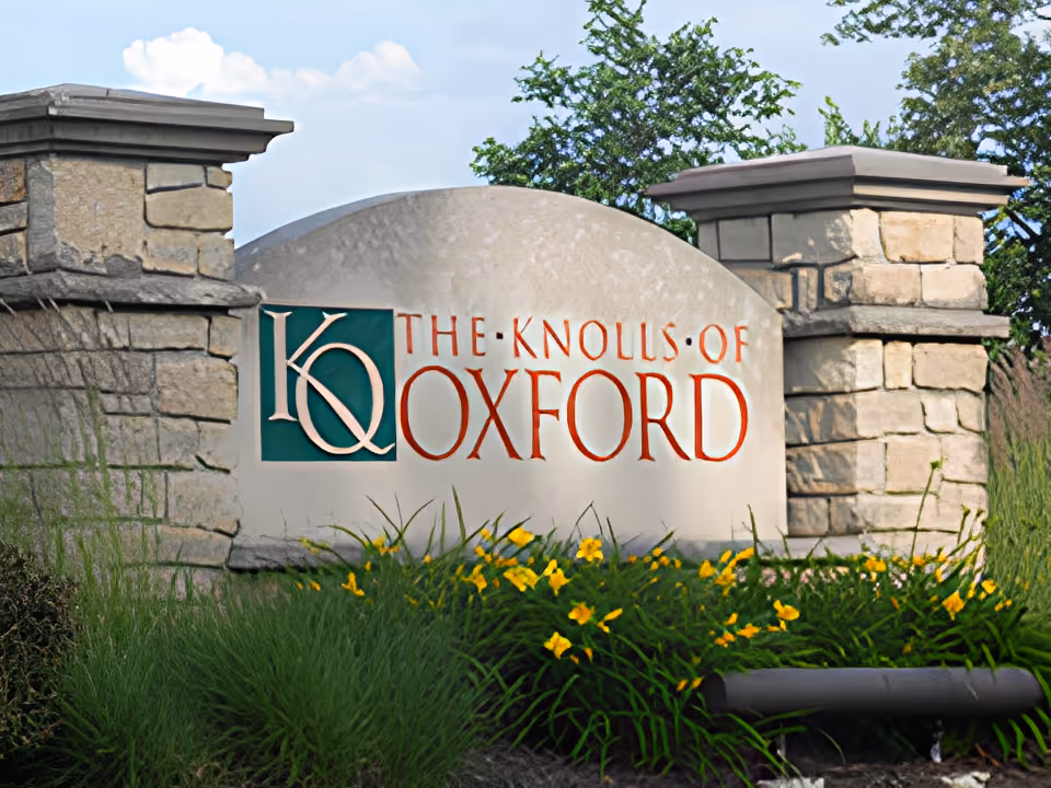 Stone entrance sign for The Knolls of Oxford surrounded by green plants and yellow flowers under a blue sky with some clouds.