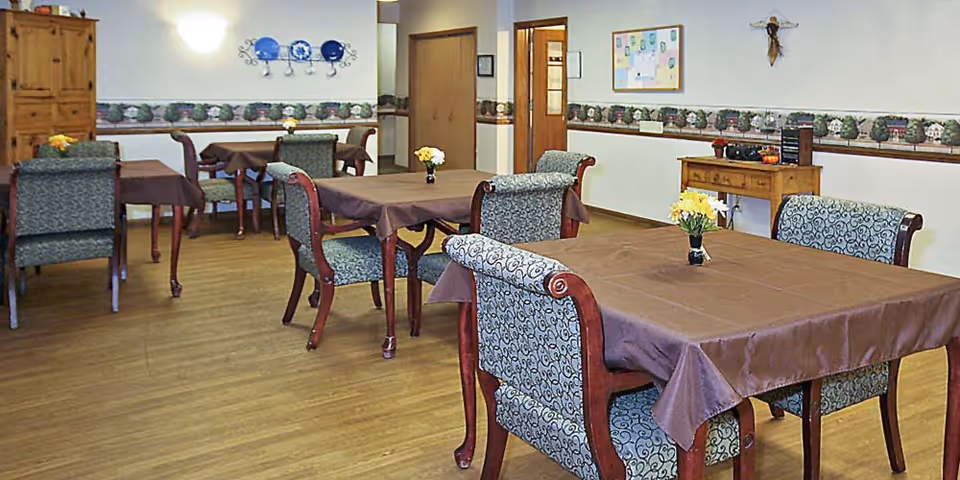 Dining room with multiple tables covered in brown tablecloths and upholstered chairs, each table topped with a small flower centerpiece and wood flooring.