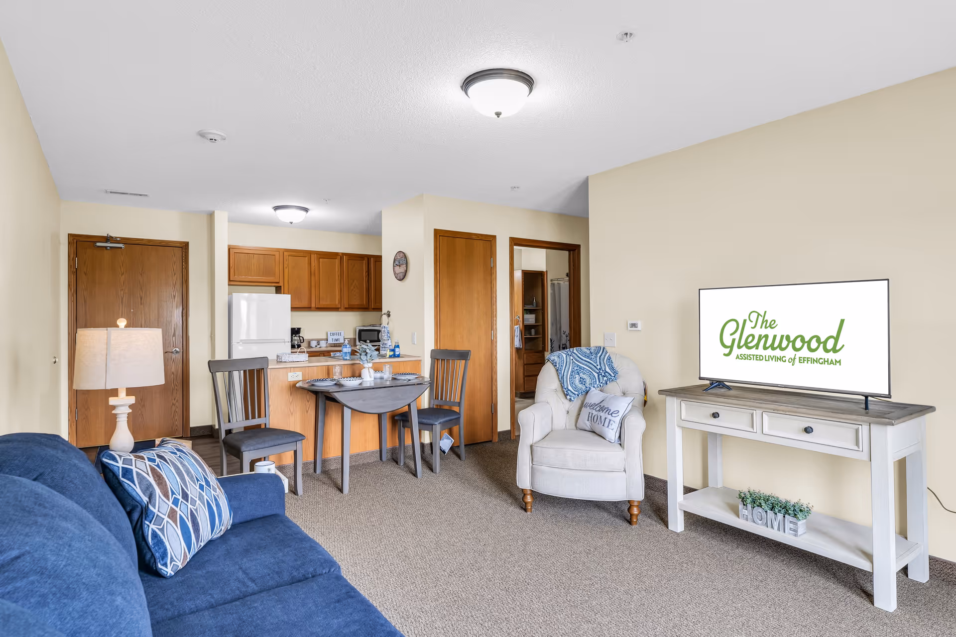 Interior view of a living area in an assisted living facility featuring a blue couch with patterned pillows, a white armchair with a 'Welcome Home' pillow and a blue throw, a small dining table with two chairs, a kitchen area with wooden cabinets and a white refrigerator, and a TV on a white console displaying 'The Glenwood Assisted Living of Effingham' logo.