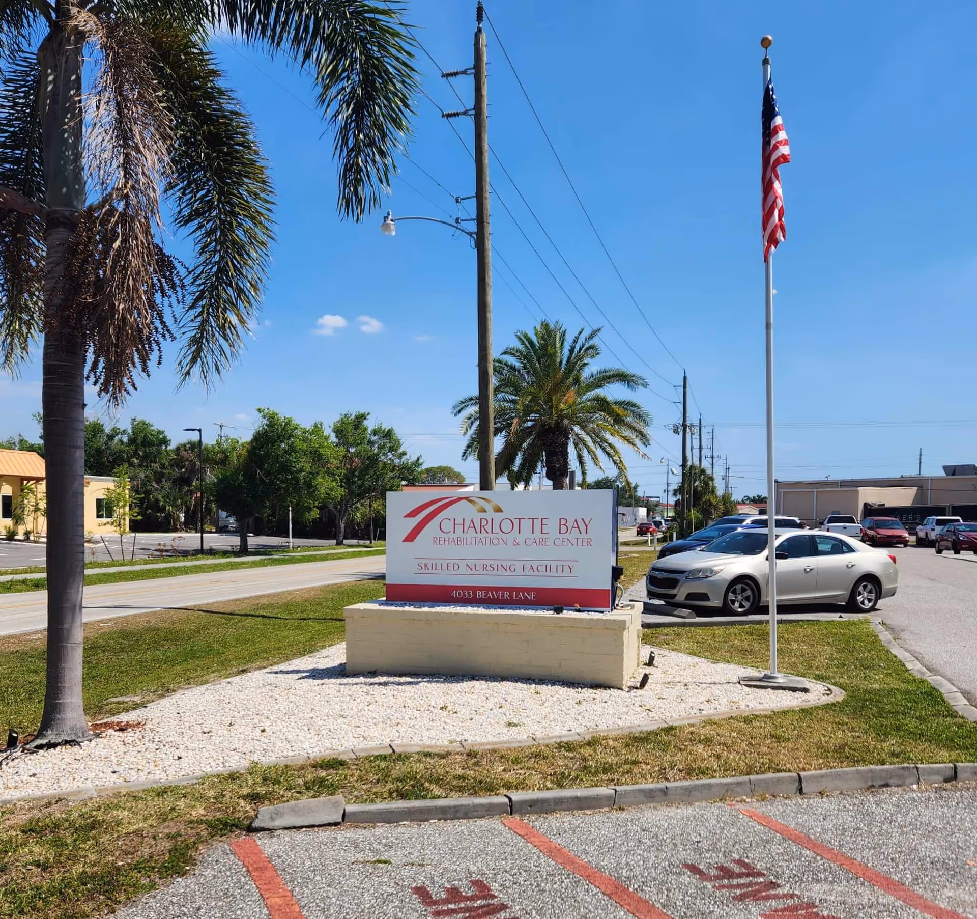 Outdoor view of the entrance area of Charlotte Bay Rehabilitation & Care Center featuring a sign with the facility's name and address, palm trees, an American flag on a flagpole, parked cars, and a clear blue sky.
