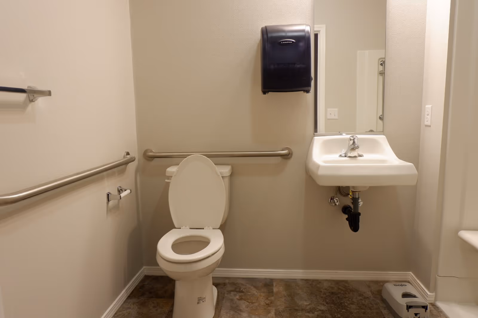 A clean and accessible bathroom with a white toilet with the lid open, a white wall-mounted sink with a faucet, a large mirror above the sink, stainless steel grab bars on the walls, a black paper towel dispenser mounted above the toilet, and a soap dispenser on the floor to the right of the sink.