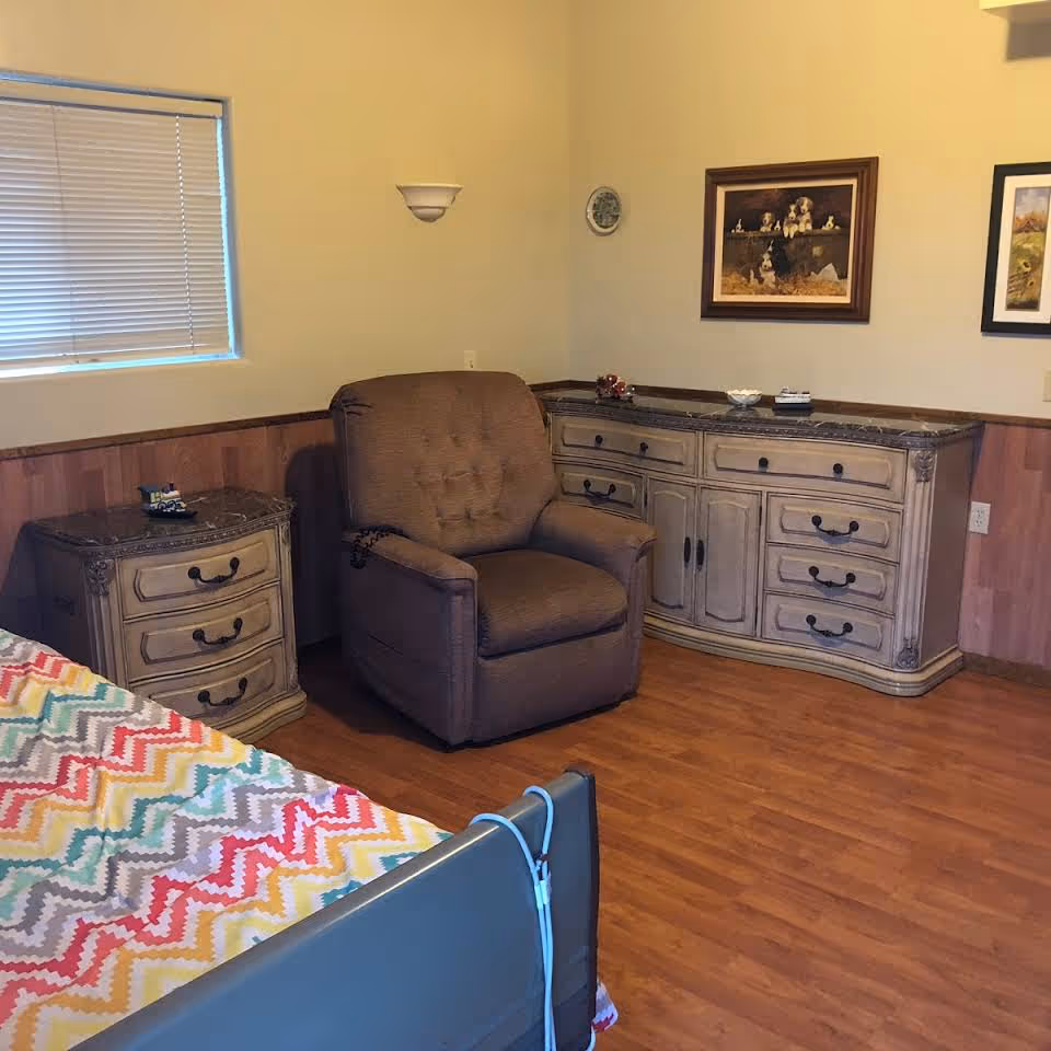 A furnished bedroom corner with a patterned bedspread, a brown recliner, and antique-style dressers along a wood-paneled wall.