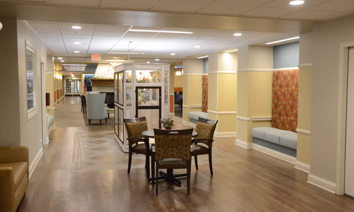 Interior hallway of a senior living facility with wooden flooring, beige walls, and recessed lighting. There is a small round table with four patterned chairs in the center, built-in cushioned benches with decorative panels along the right wall, and a glass display case near the middle. The hallway extends into the distance with additional seating and an exit sign visible.