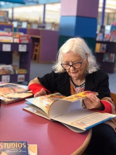 An elderly woman with white hair and glasses is sitting at a round table in a library or reading area, looking through a large colorful book. Bookshelves filled with books are visible in the background.