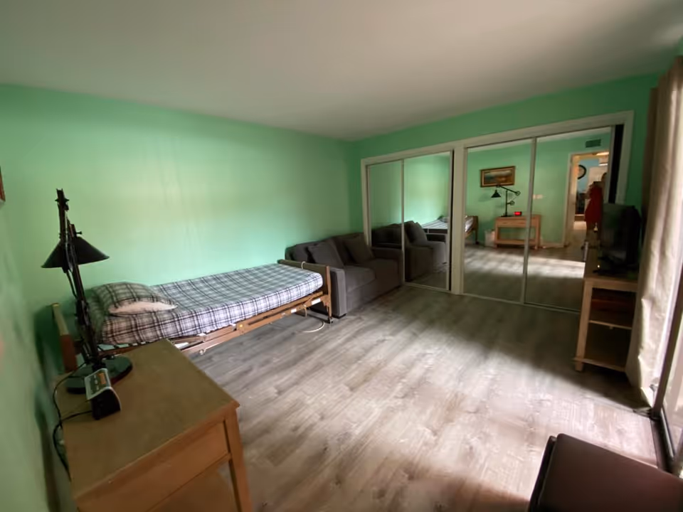A bedroom with light green walls and wood flooring. The room contains a single bed with a plaid blanket, a small gray sofa, a wooden desk with a lamp and clock, and a TV on a wooden stand. There is a large mirrored closet on one wall and a window with curtains letting in natural light.