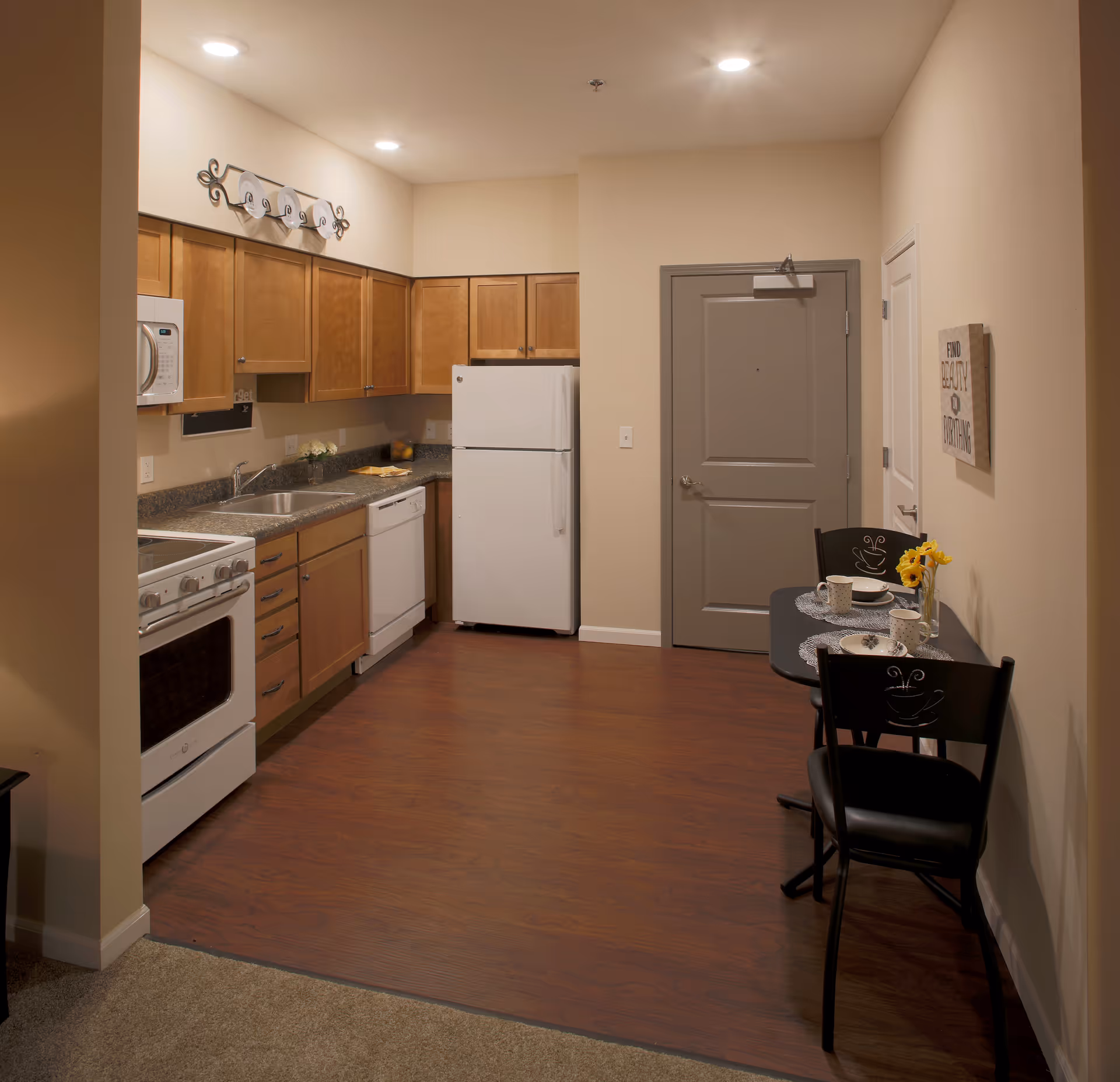A compact kitchen area with wooden cabinets, a white refrigerator, stove, microwave, and dishwasher. There is a small dining table with two chairs, set with plates, cups, and a vase with yellow flowers. The floor is a combination of wood and carpet, and the walls are painted beige with a gray door in the background.