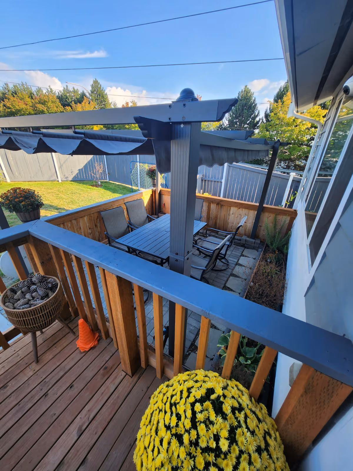 Outdoor patio area with a wooden deck and railing, featuring a table with four chairs under a pergola with a retractable canopy. There are potted plants including a large pot of yellow flowers and a basket filled with pine cones. The patio is adjacent to a grassy backyard enclosed by a gray fence, with trees and a clear blue sky in the background.