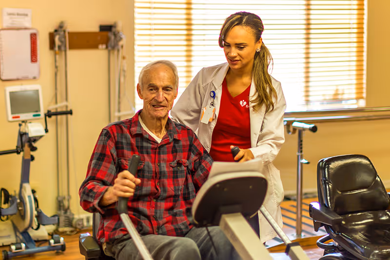 An elderly man in a red plaid shirt is using a seated exercise machine while a female healthcare worker in a white coat and red shirt assists him in a rehabilitation center room with exercise equipment and wooden blinds on the windows.
