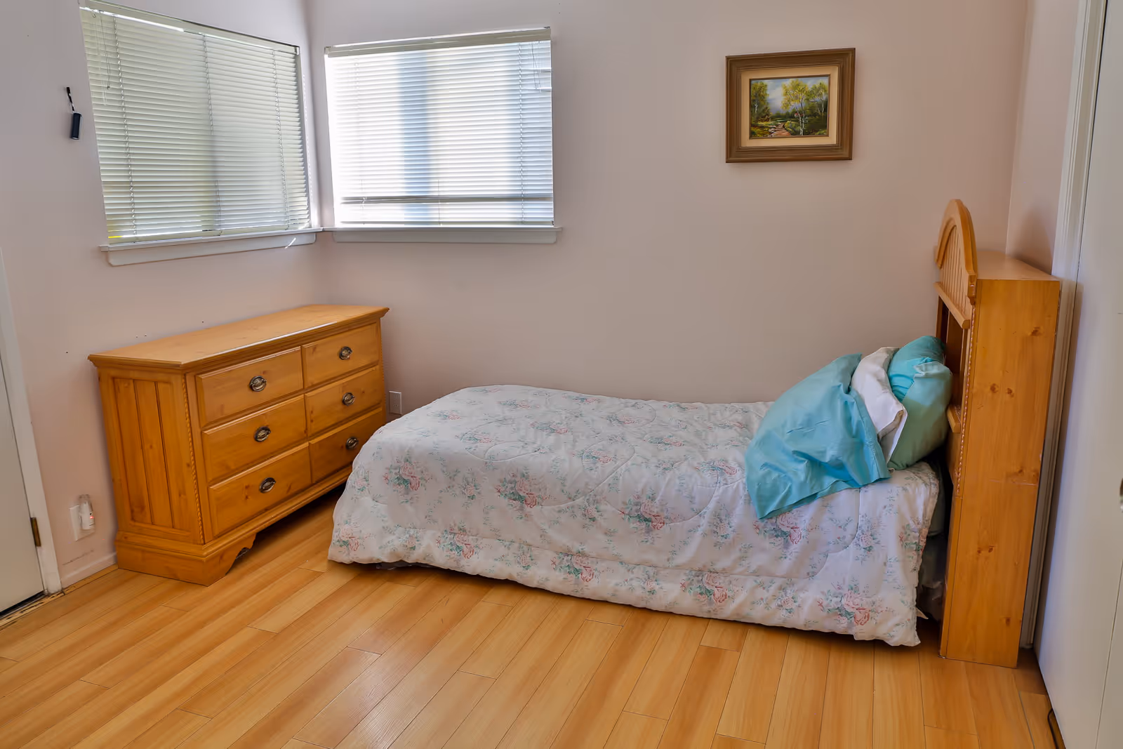 A small bedroom with a single bed covered in a floral quilt and blue pillows. There is a wooden dresser with six drawers against the wall under two windows with closed blinds. A framed landscape painting hangs on the wall above the bed. The floor is light wood.