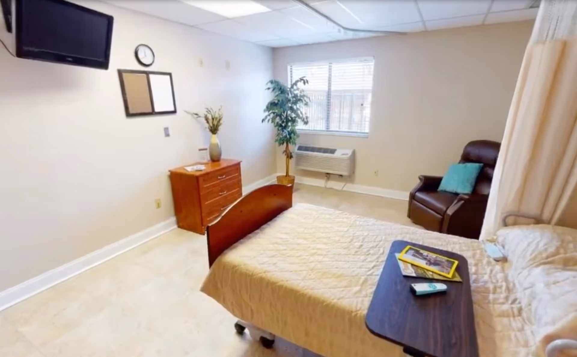 A senior living facility bedroom with a bed covered in a beige quilt, a small wooden dresser with a vase of flowers, a wall-mounted TV, a clock, a window with blinds, a potted plant, and a brown recliner chair with a teal pillow.