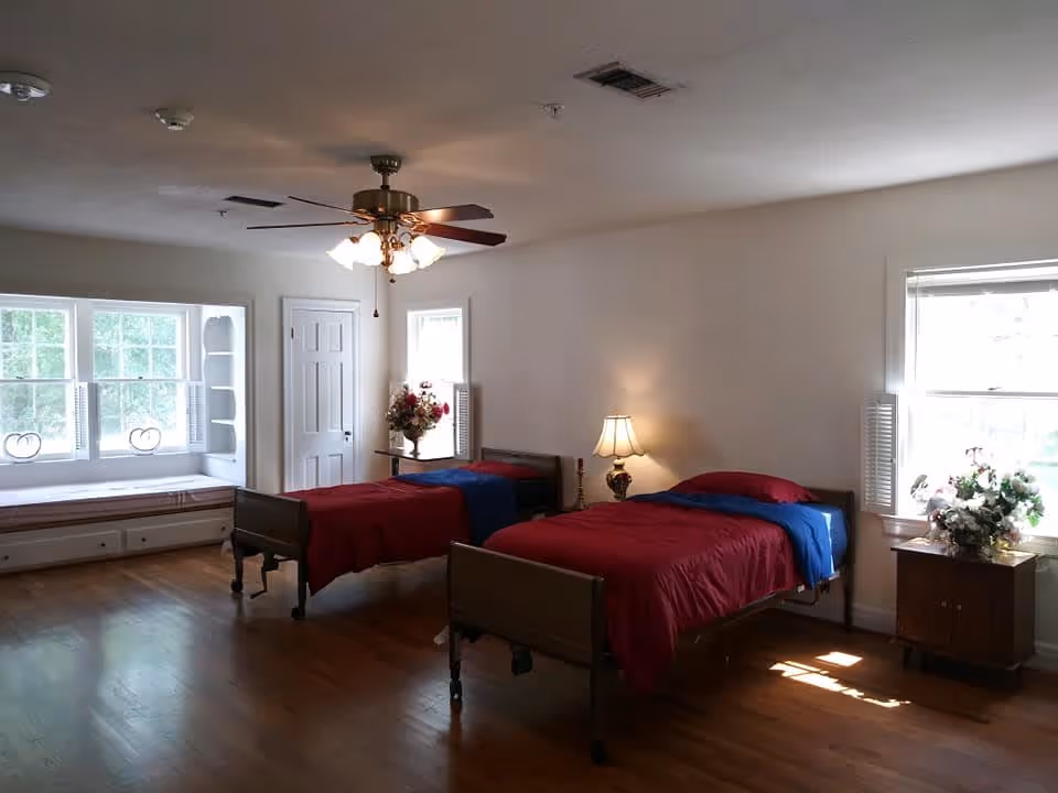 A bright bedroom with two single beds covered in red and blue bedding. There is a wooden nightstand with a lamp and a flower arrangement between the beds. The room has hardwood floors, white walls, a ceiling fan with lights, and multiple windows allowing natural light to enter. A built-in window seat with cushions is visible on the left side.