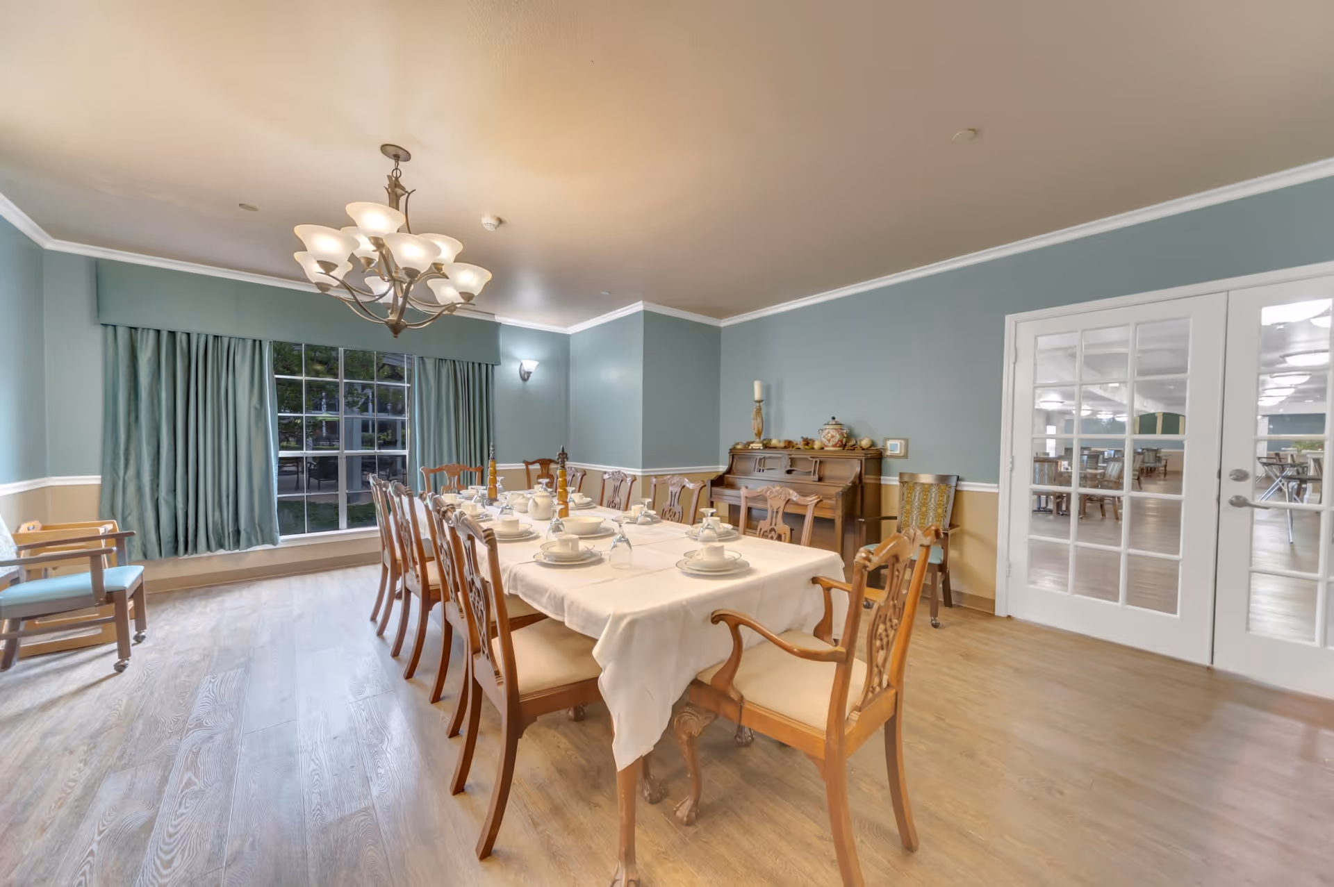 A formal dining room with a long table covered with a white tablecloth, set with plates, bowls, glasses, and candles. The room has wooden chairs with cushions, a large window with green curtains, a chandelier overhead, and a piano against the wall. French doors lead to another room with more tables and chairs.