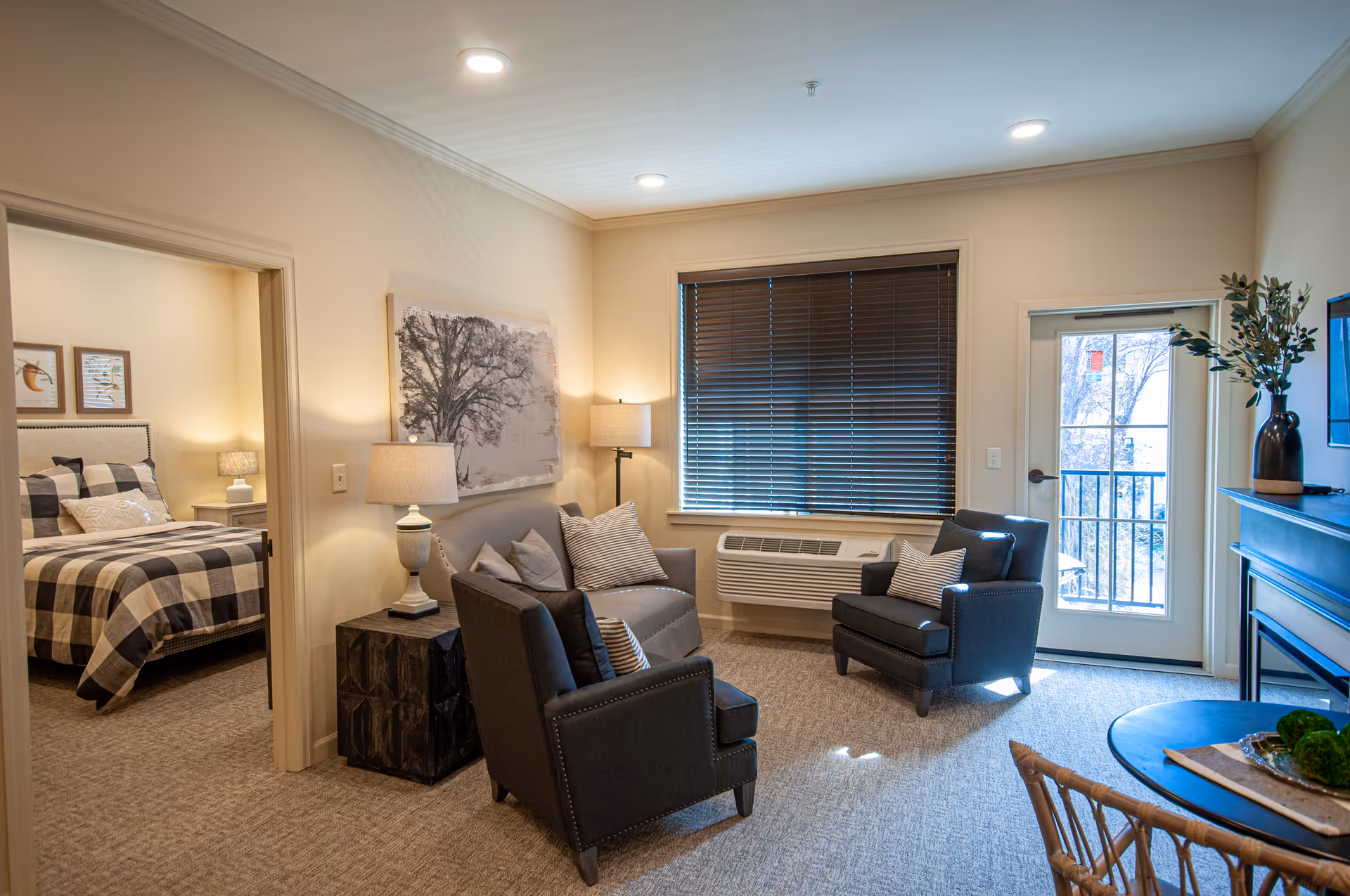 A cozy living room in a senior assisted living facility featuring two dark armchairs with striped pillows, a small gray sofa with multiple pillows, a side table with a lamp, and a large window with closed blinds. To the right, there is a door leading outside with a small balcony visible. A round table with a decorative centerpiece and a TV mounted above a fireplace are also present. In the background, a bedroom with a checkered bedspread and bedside lamps is visible through an open doorway.